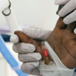 A nurse’s gloved hand holds a patient’s hand during a blood oxygen check.