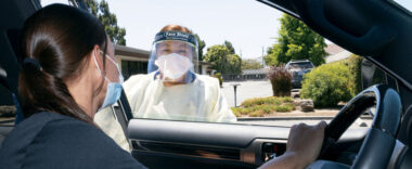 A healthcare provider in full PPE speaks to a patient with COVID-19 through a car window.